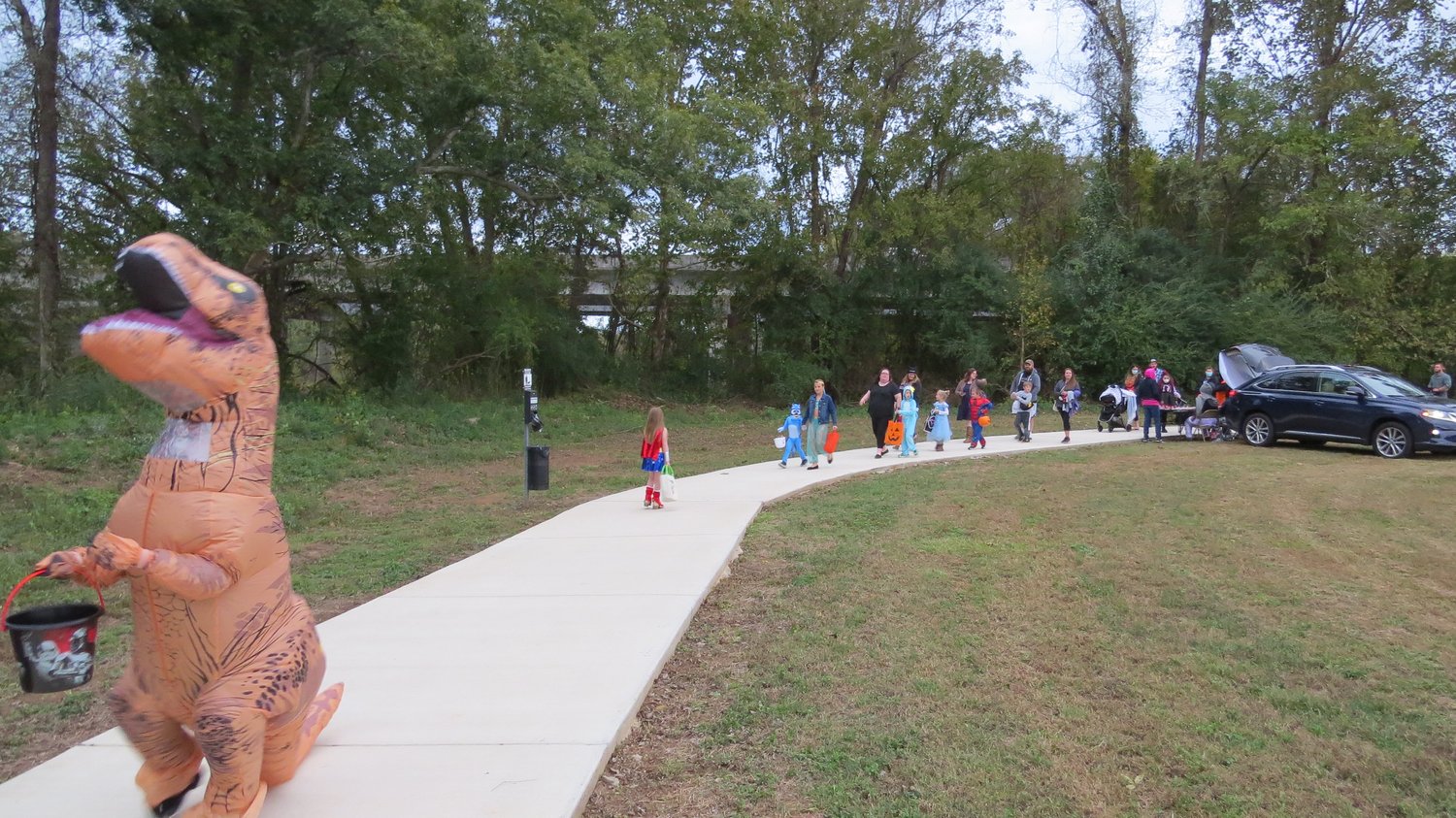 veteranshalloween1 A parade of small children in Halloween Costumes getting out of cars going trick or treating. An adult sized dinosaur is in the foreground trick-or-treating at the Veterans Park Loop Trail