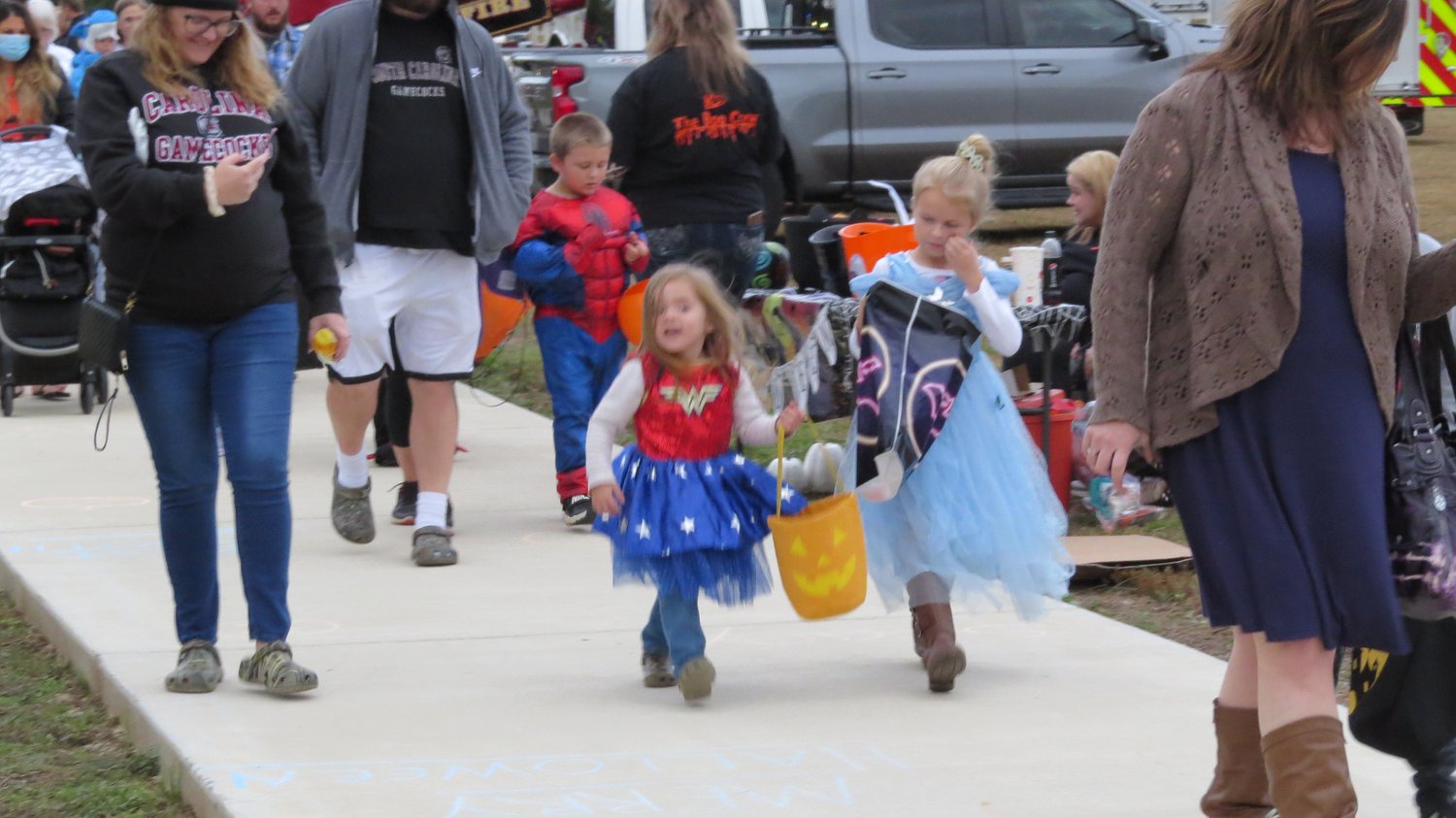 veteranshalloween3 Two small children one with a wonder woman costume and another witha princess costume trick or treating with a small boy with a spider man costume on surrounded by adults at Veterans Park Loop Trail
